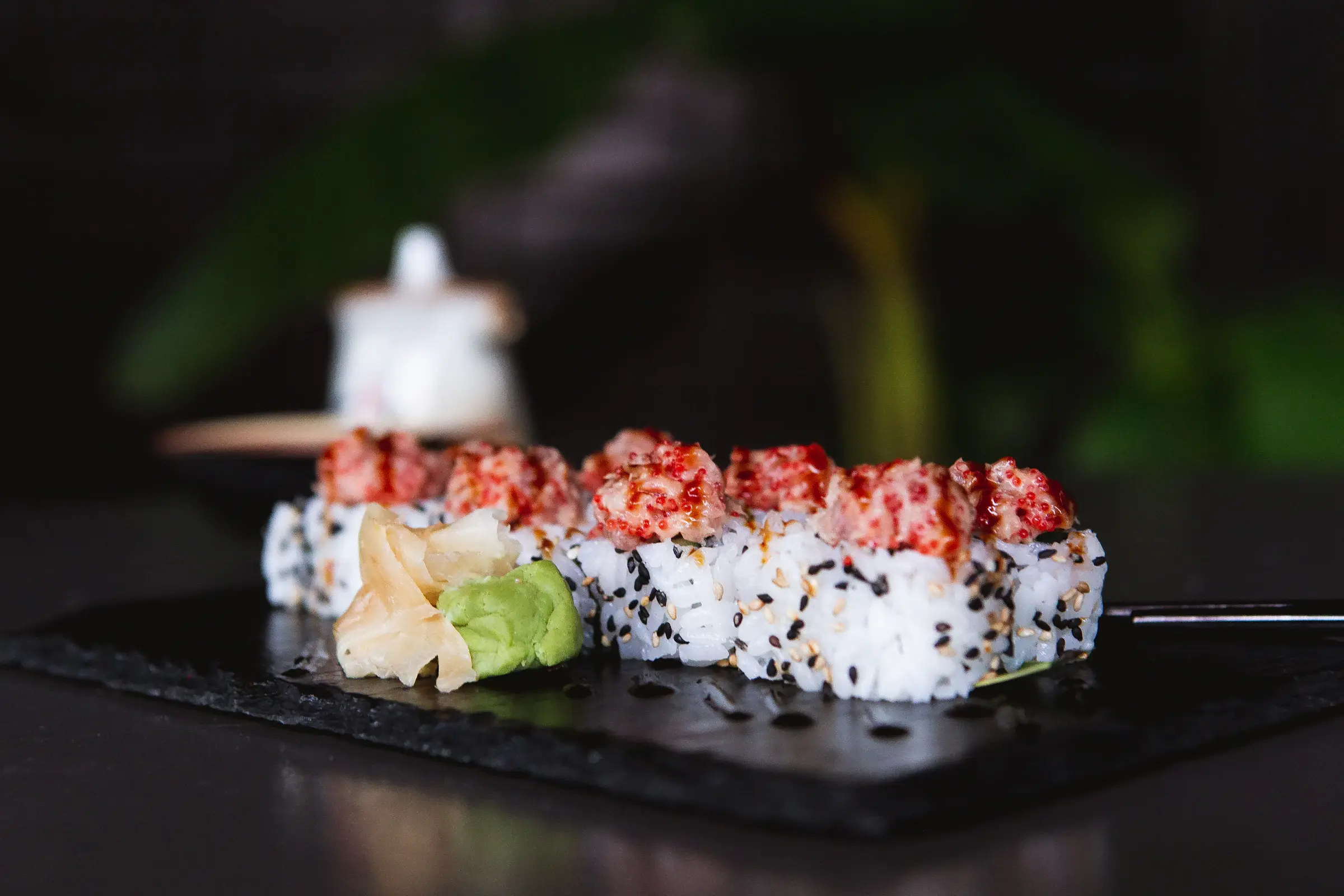 Rollos de atún picante cubiertos de ajonjolí blanco y negro sobre una tabla de pizarra, con jengibre encurtido, wasabi y una jarrita de salsa de soya al fondo